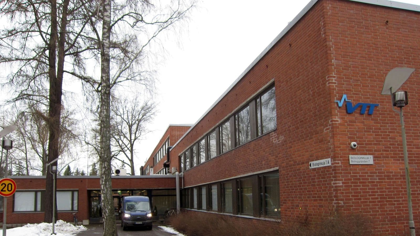 VTT:n punatiilinen toimitalo Biologinkuja 7:ssä Espoossa, edessä puita ja sinertävä pakettiauto