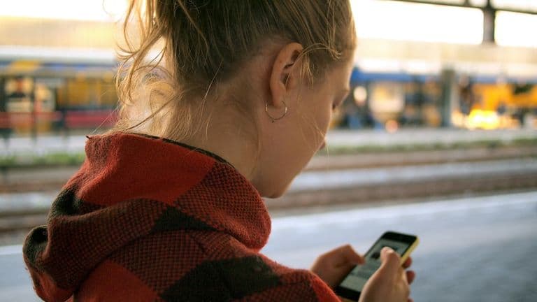 Nuori punaruutuisessa huppuhuppatakissa selaa älypuhelinta rautatieaseman laiturilla