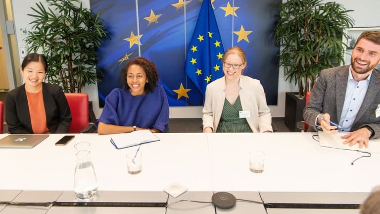 Neljä hymyilevää henkilöä istuu kokouspöydän ääressä EU-lipun edessä Euroopan komission tilaisuudessa