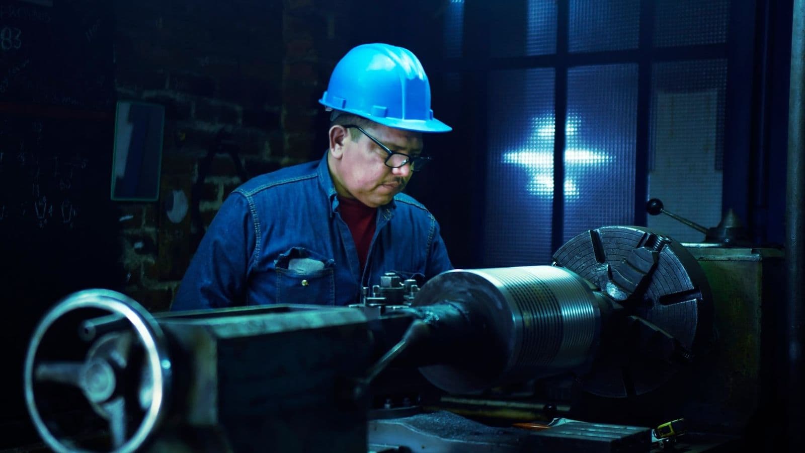 Kypäräpäinen metallityöntekijä seisoo sorvin ääressä hämärässä konepajassa ja tarkkailee työstettävää kappaletta