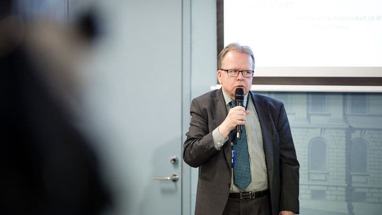 Silmälasipäinen, tummapukuinen mies puhuu mikrofoniin tilaisuudessa, taustalla esityskalvo ja vaalea ovi