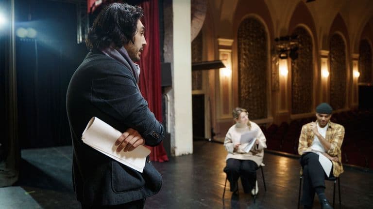 Ohjaaja seisoo näyttämöllä käsikirjoitus kainalossaan ja katsoo kahta istuvaa näyttelijää vanhassa teatterisalissa