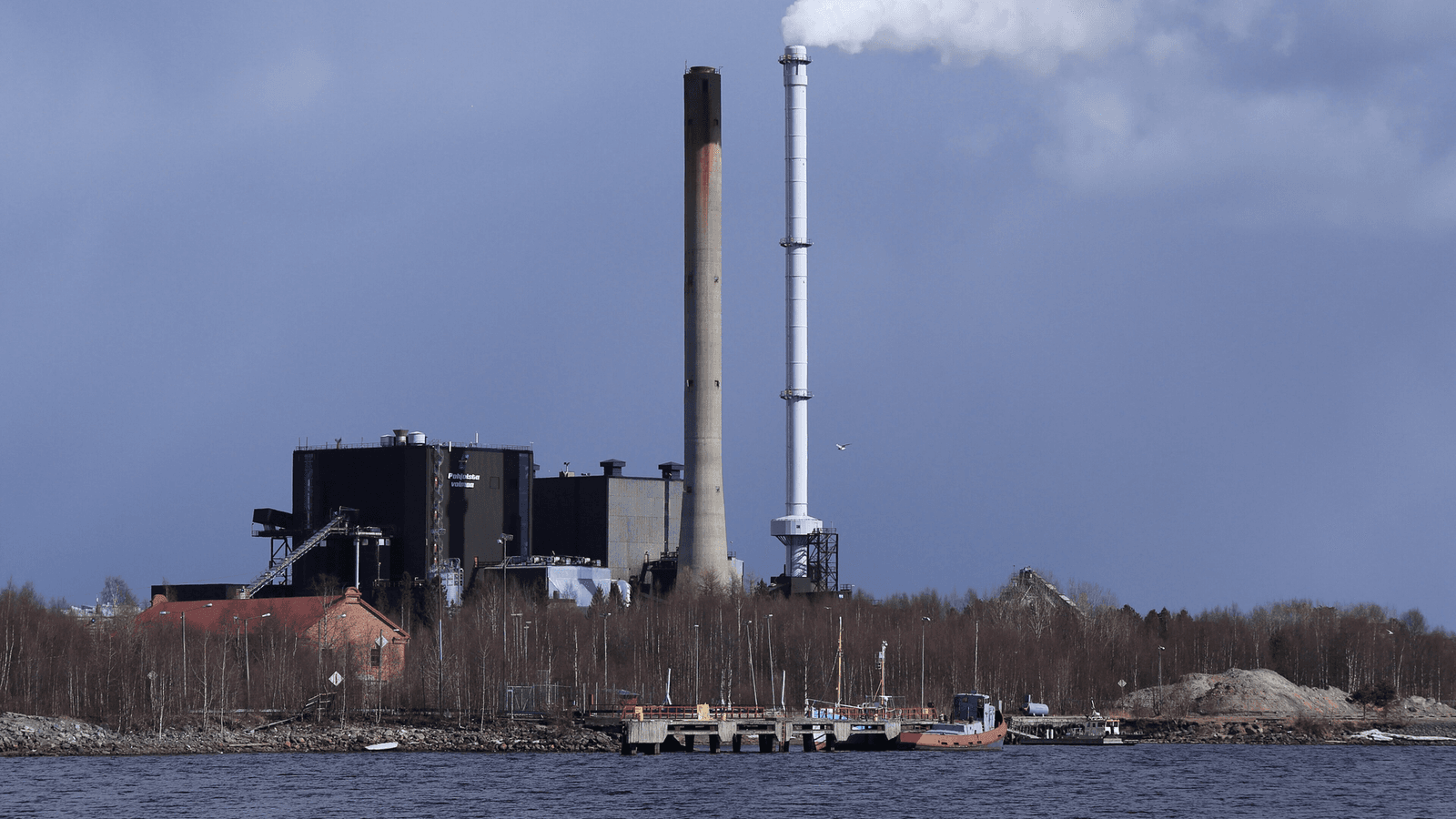 Toppilan voimalaitos Oulussa meren takaa kuvattuna, kaksi korkeaa savupiippua ja tumma voimalarakennus.