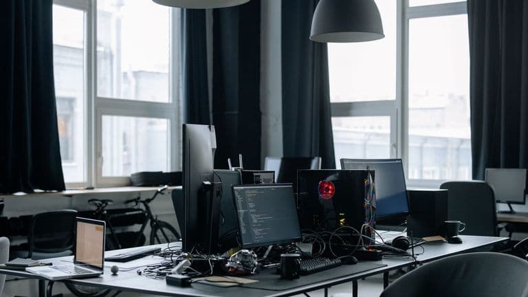 Ohjelmistokehittäjän työpöytä, jolla on useita näyttöjä koodineditorin kanssa, tietokone ja kannettava tilavassa toimistossa.